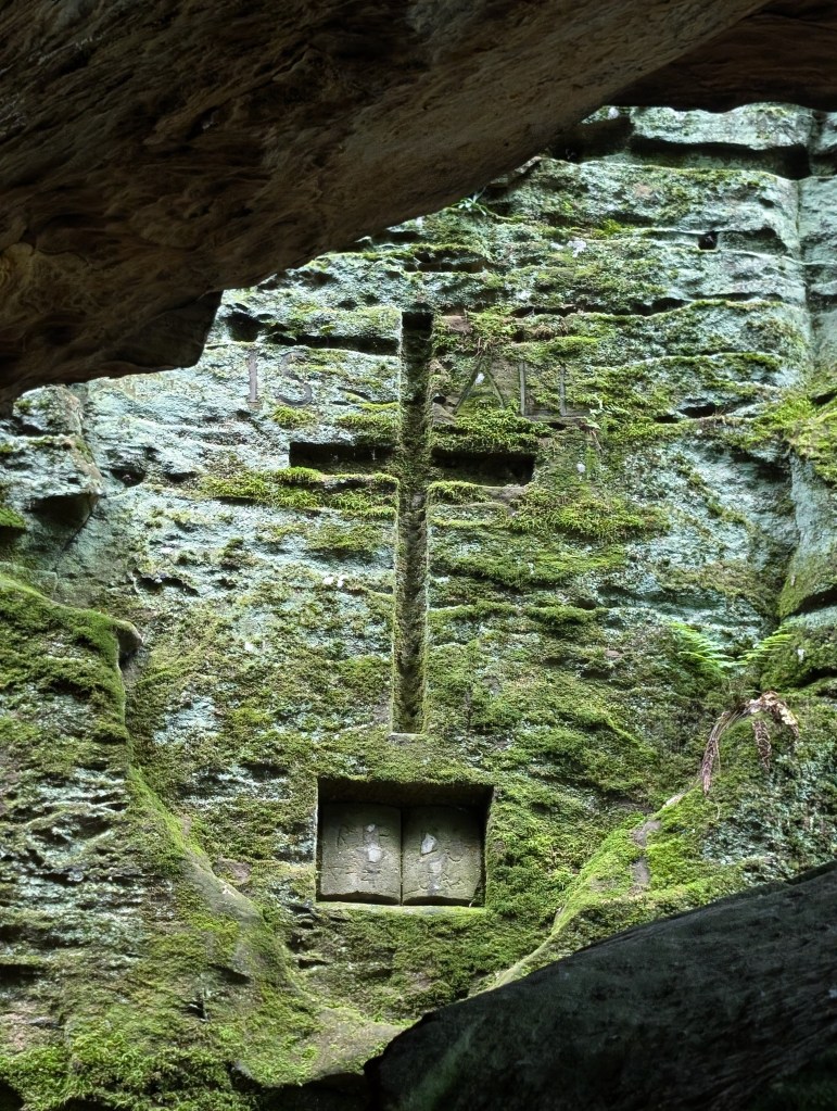 A large cross carved into the stone with a Bible carved out beneath it. There also appears to be words IS ALL carved near the cross. The whole thing is covered over with moss.