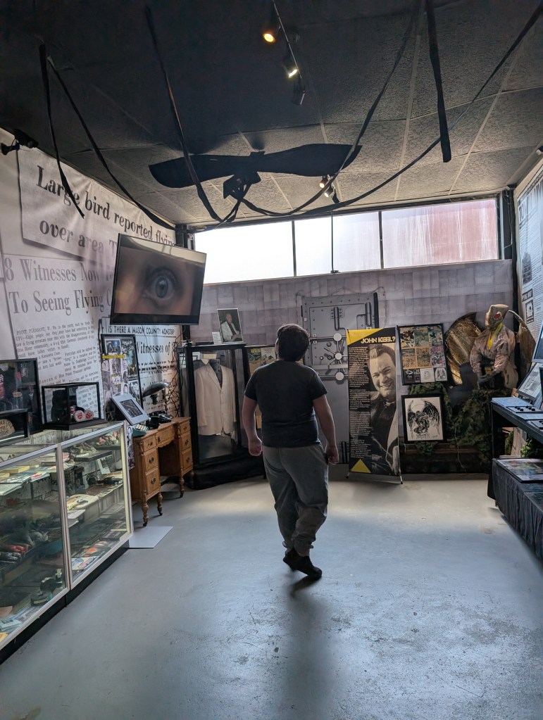 A teen boy in sweats and a tshirt walks around a large open room. His back is to us. There is a TV on the wall with an open eye and several cases of artifacts related to the Mothman case.