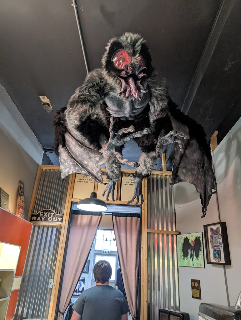 A hanging model where Mothman looks very furry with an open mouth that splits in six parts. A teen boy walks under it, his back to us.
