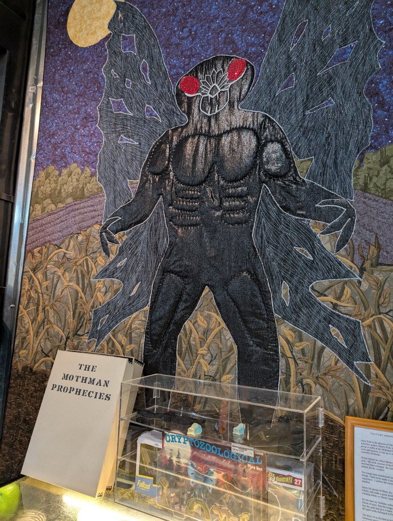 A large quilt with a shiny black Mothman embroidered on it with bright red eyes and tattered wings.
