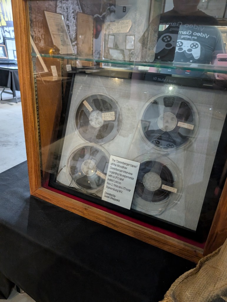 A glass case contains four reel to reel wheels. A sign says "The 'Derenberger taps' of the Woodrow Derenberger interview regarding his experience with a UFO and Indrid Cold November 2, 1966 on I-77 near Parkersburg, WV"