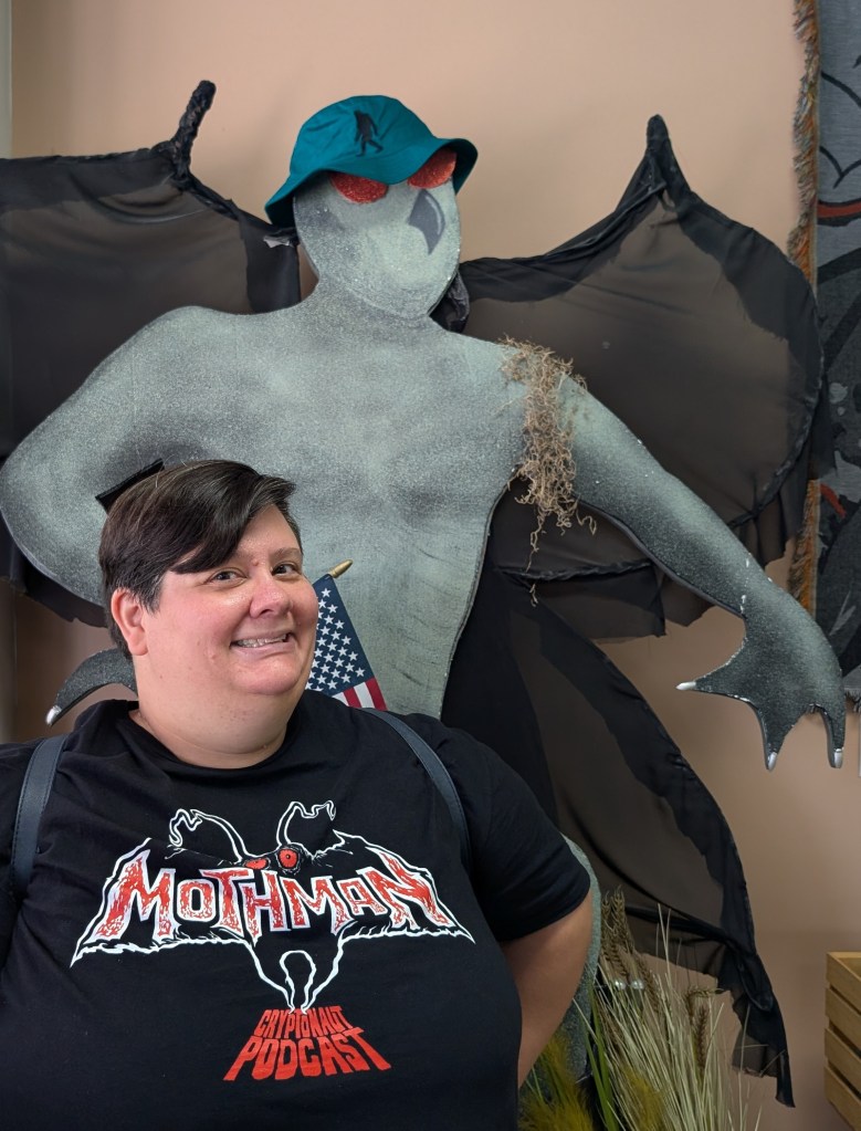 A round-faced person with short brown hair is wearing a black t-shirt that has Mothman written in red with Cryptonaut Podcast beneath it. They stand in front of a large grayish green figure with black wings, a grey beak, red eyes, and a blue bucket had.