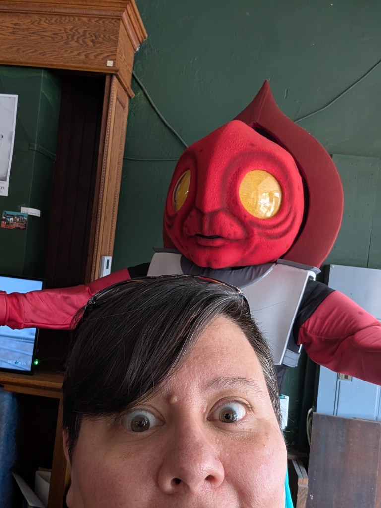 A round-faced person with short brown hair is taking a selfie with a tall alien figure. It has red arms and a large red face with a red spade-shaped hood behind it. It has large yellow eyes.