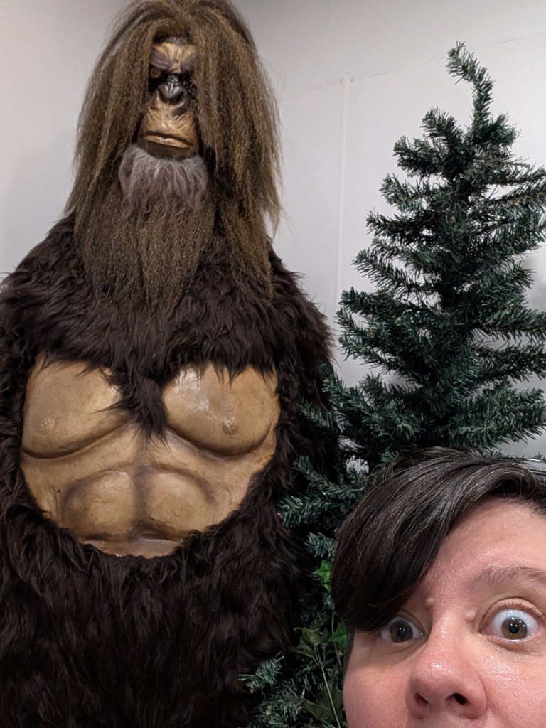 A round-faced person with short brown hair takes a selfie with a fake pine tree and a large Bigfoot figure that has a bare chest, a coat of shaggy fur, and crimped brown hair for some reason.