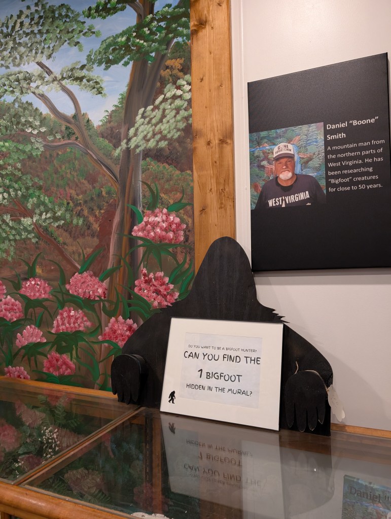 Half of a Bigfoot cut-out holds a sign that says "Can you find the 1 Bigfoot hidden in the mural?"