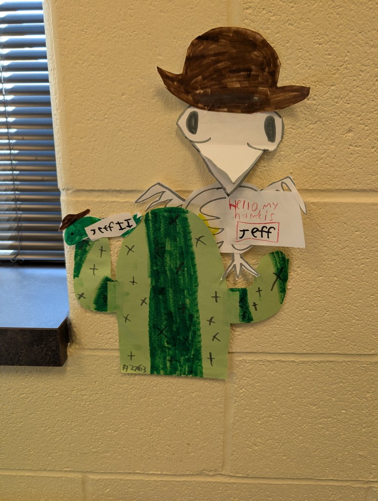 A hand-drawn and cut out bird skeleton wearing a bowler hat and a name tag that says "Hello, my name is Jeff". It is taped on a wall behind a hand-drawn green cactus. On the cactus is a small hand-drawn snake with a tiny cowboy hat and a nametag that says "Jeff II".