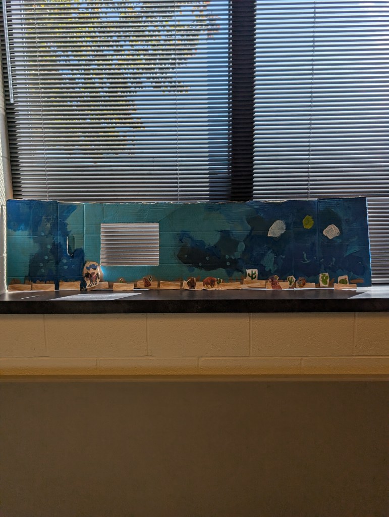 A desert scene is painted by a young kid on a long piece of scrap cardboard. The sky is various colors of blue. There are tiny cut out pieces of desert along the bottom and cut out animal figures glued on.