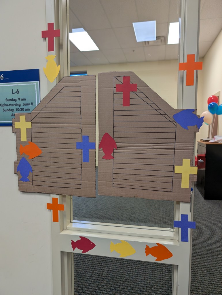 A pair of saloon doors drawn on cut pieces of cardboard. A bunch of fish and cross shapes in a variety of colors are glued around the saloon doors and the door frame.