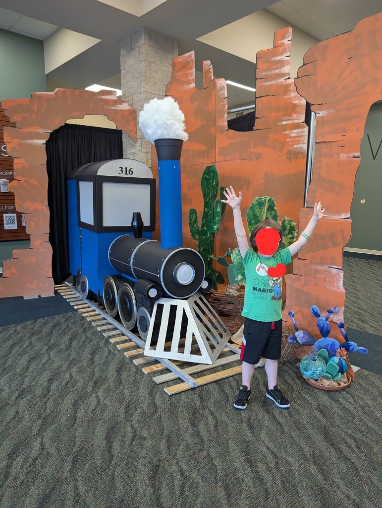 An elementary-aged child is standing with their arms outstretched as if afraid. Their face is obscured. Behind them is a very impressive train prop surrounded by prop canyons and cactuses.