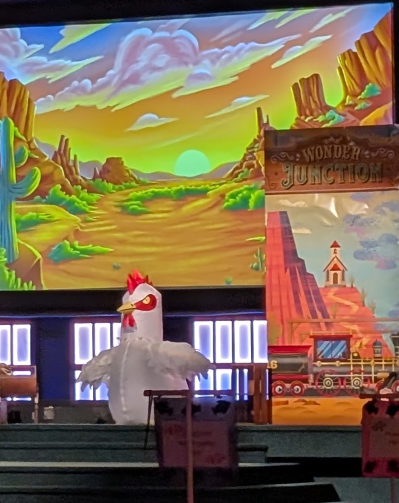 A person in a giant inflatable chicken costume sits on a stage. There is a big screen behind them with a painted western scene and a banner beside them that says "Wonder Junction" and shows a train in a desert scene.