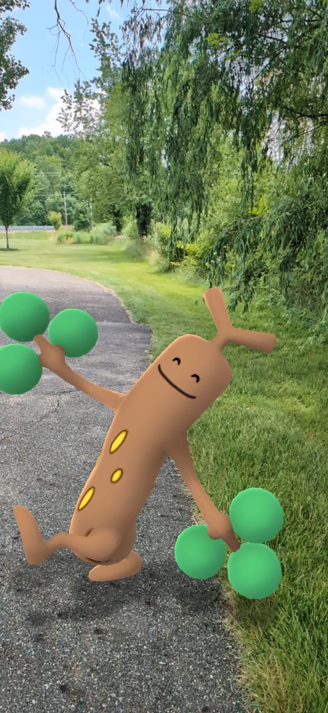 A sudowoodo (tree-like Pokemon) strikes a happy pose on a paved path with green grass and trees in the background.
