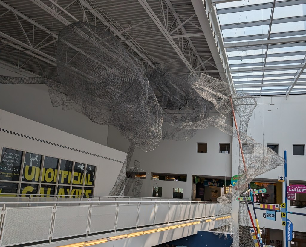 A very large wire model of a human skeleton hangs from the ceiling of the second floor of an open museum. 