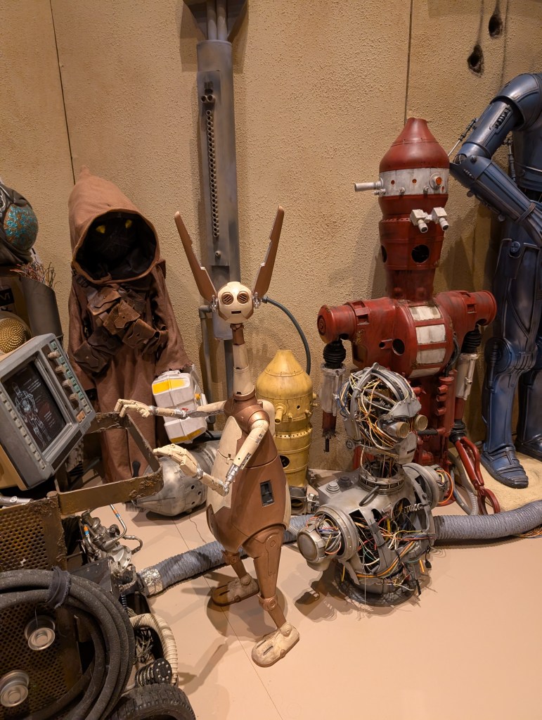 A collection of different droids from Star Wars, including the torso and head of an unfinished humanoid droid, a red and white droid that looks sort of like a fire hydrant, and a little guy with big ears that is pushing a junk cart. 