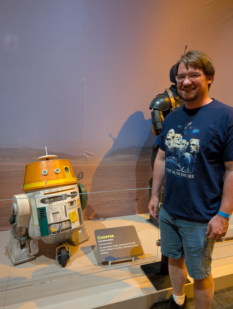 A man with blond hair and a happy smile stands next next to a small droid with an orange top and green and yellow highlights. The droid looks innocent but he is a menace.