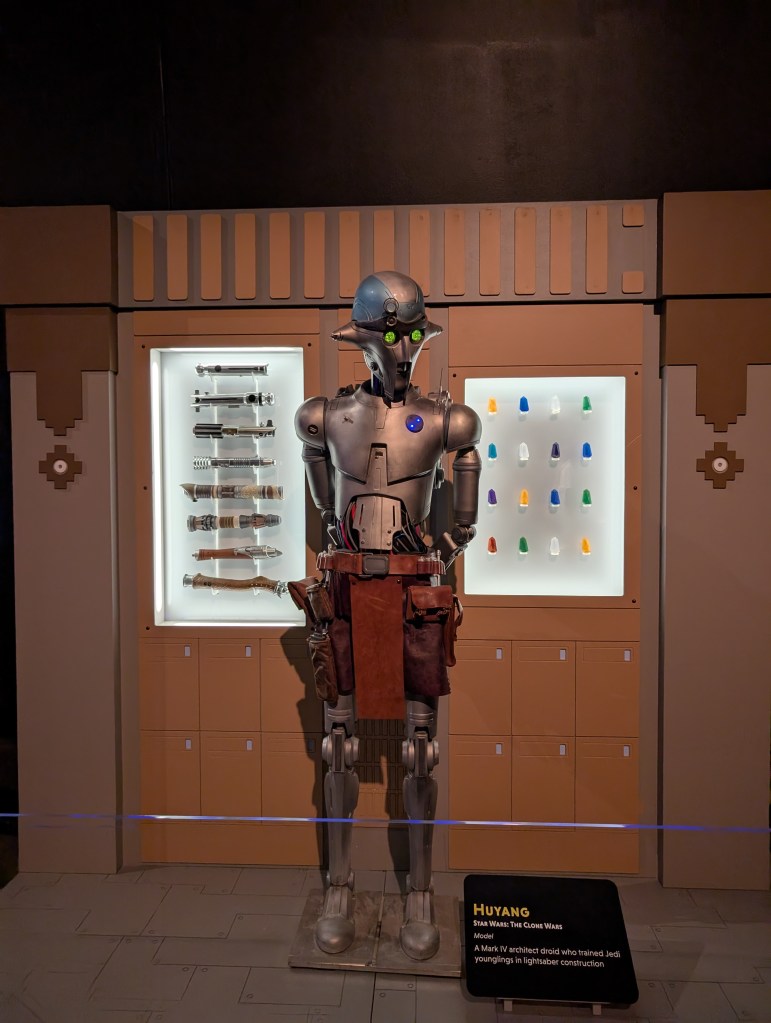 A tall droid, shiny copper colored, with a leather belt and many hanging pockets. A sign identifies him as Huyang.
