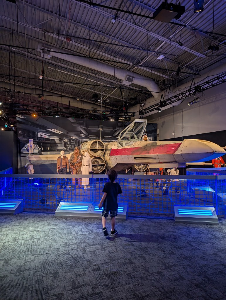 A young boy faces away from the camera, looking at a large, sleek, white spaceship with a red streak on the side, clearly to make it go faster. 