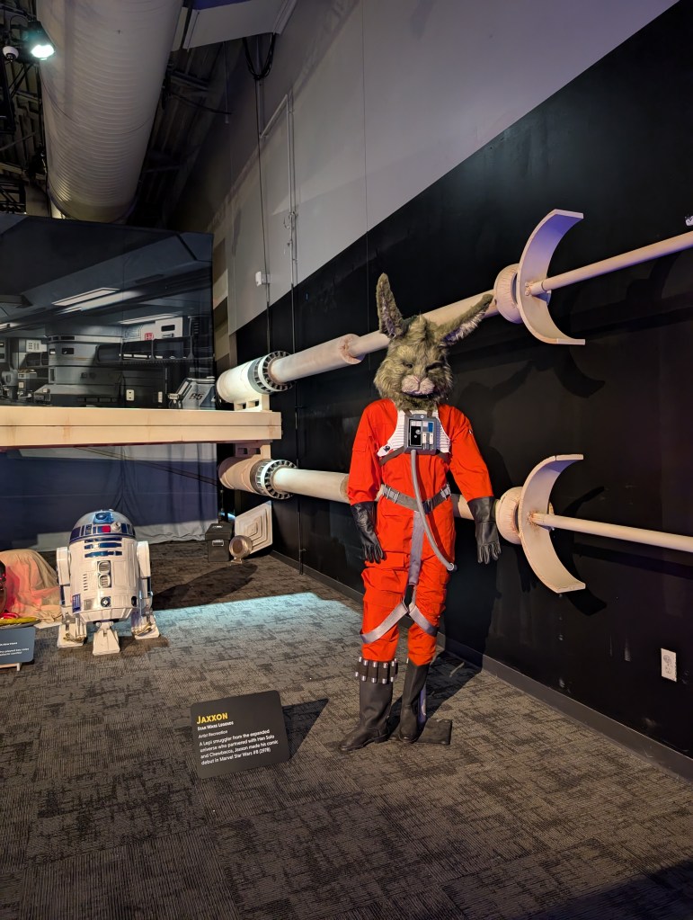 In the background, an iconic droid in blue and white looks at the picture taker. In the foreground is a large humanoid rabbit in a bright orange flight suit. A sign identifies him as Jaxxon.