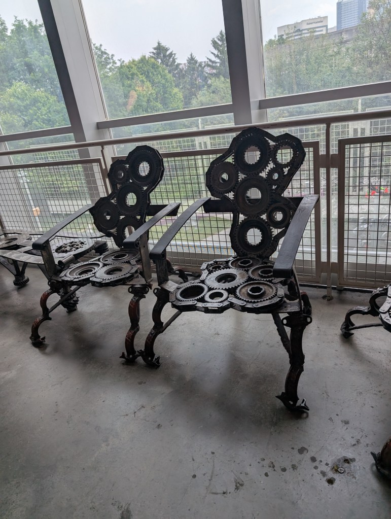 Two chairs that appear to b e made out of metal gears, wheels, and chains melded together.