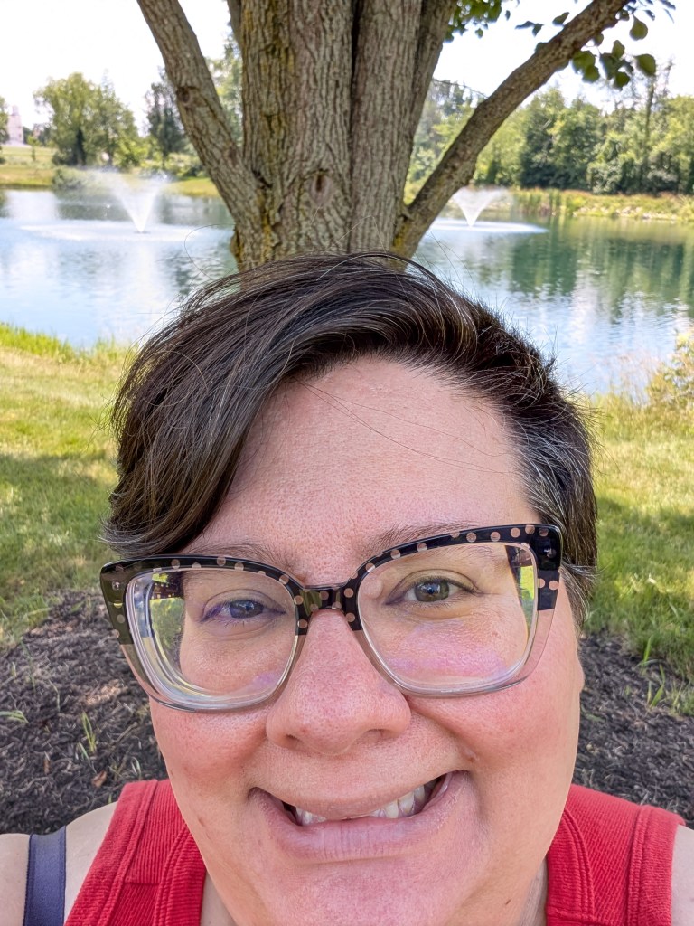 A round-faced person with short brown hair and big glasses gives an awkward smile in front of a tree that is sort of but not really positioned to show the two fountains in the lake.
