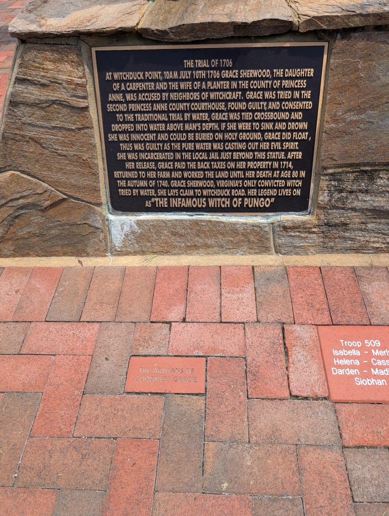 A historical marker that is set on the base of a statue. It is surrounded by a brick walkway.