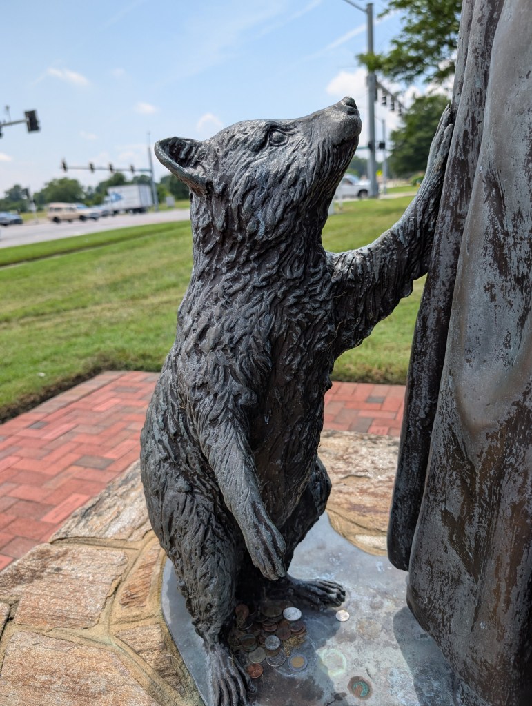 An absolutely adorable raccoon statue. It has one paw on a woman's dress and is looking up at her fondly. There are coins scattered around its feet that have presumably been placed there by other visitors.