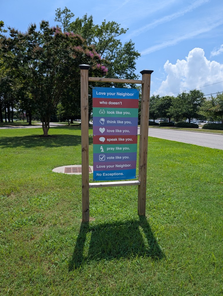 A sign in the churchyard that reads "Love your neighbor who doesn't look like you, think like you, love like you, speak like you, pray like you, vote like you. Love your Neighbor. No Exceptions."