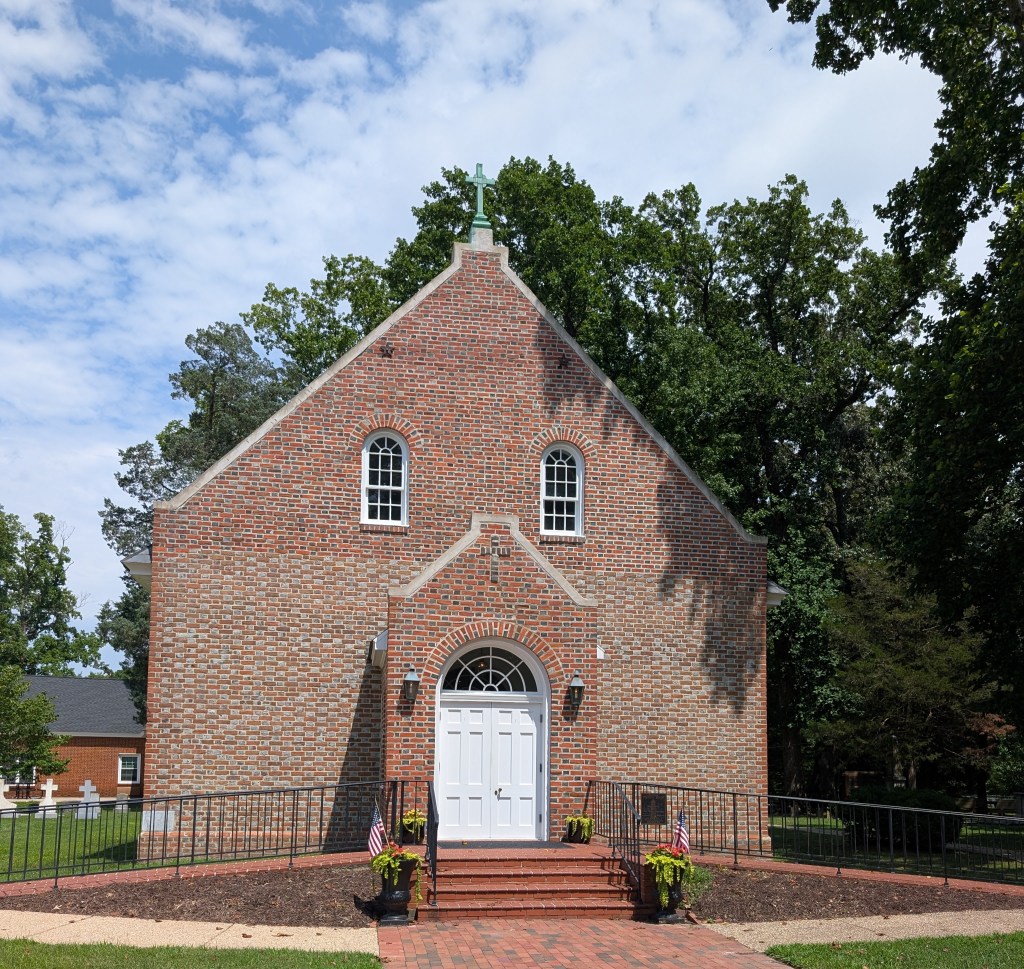 The front of a nice brick church in an old style. It has bright white double doors and two arched white windows. There is a bronze cross on top.
