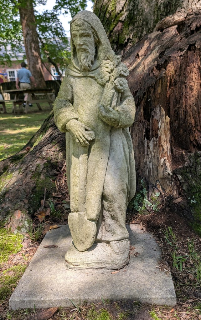A smallish, slightly worn statue of an old man with a beard in a monk's habit holding a shove with one hand and a bundle of flowers in the other.