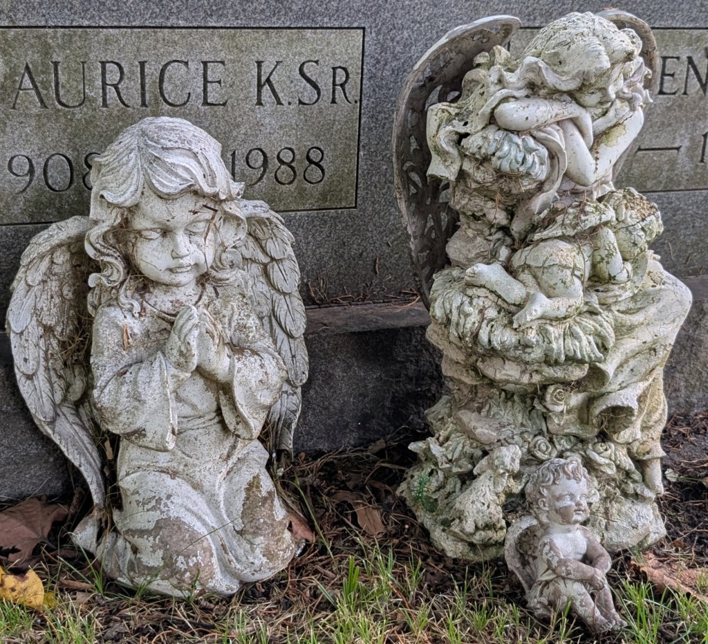 Three small worn angel statues in front of a gravestone. I do not like any of them.