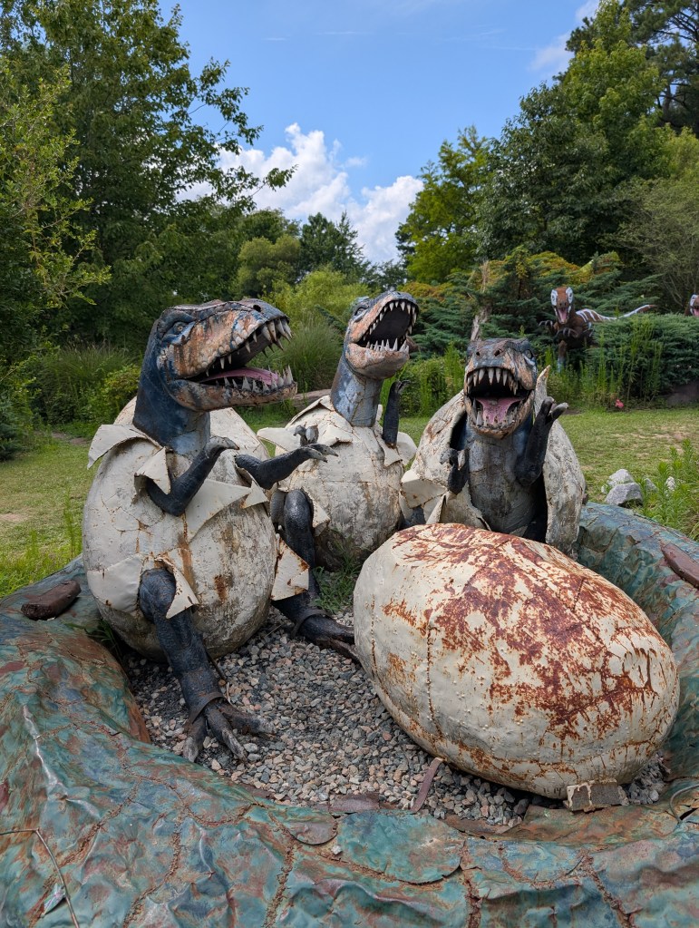 Three little metal dinosaurs are emerging from three metal dinosaur eggs. There is one unhatched egg which will probably never hatch because it is covered with the rust. They are all in a large metal nest.