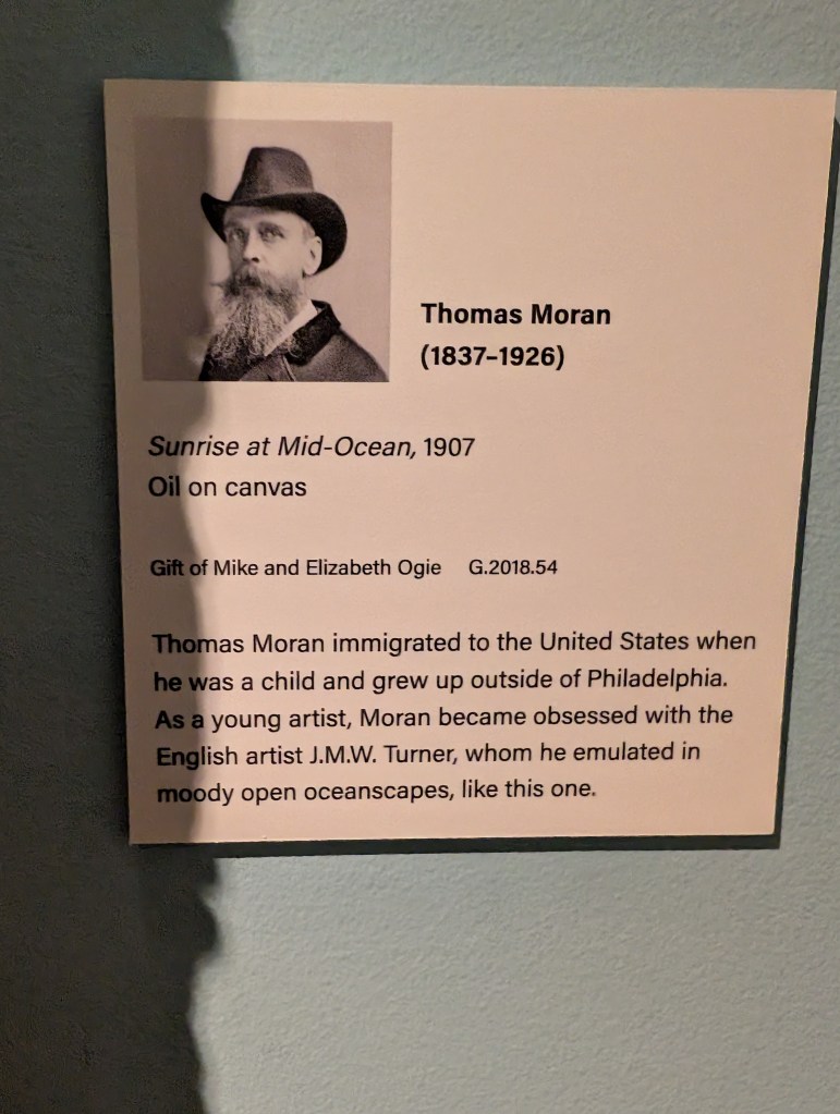 A plaque that says the art is called "Sunrise at Mid-Ocean" and was made in 1907 by Thomas Moran
