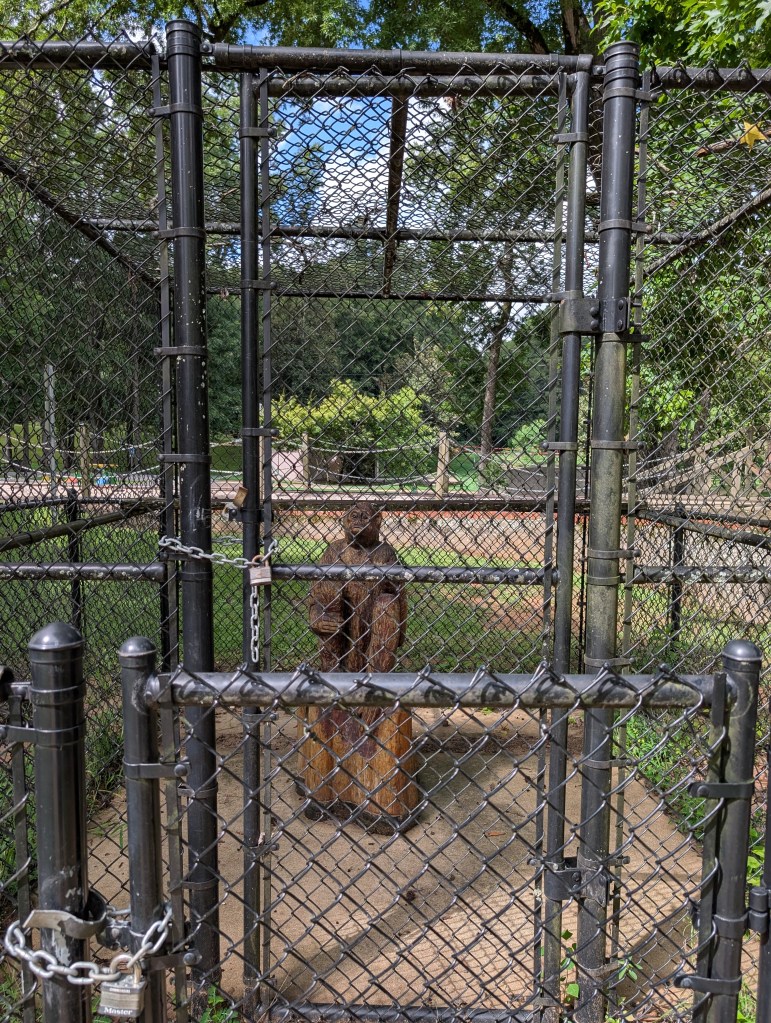 A carved wooden monkey statue is in a padlocked cage that is behind a padlocked fence.