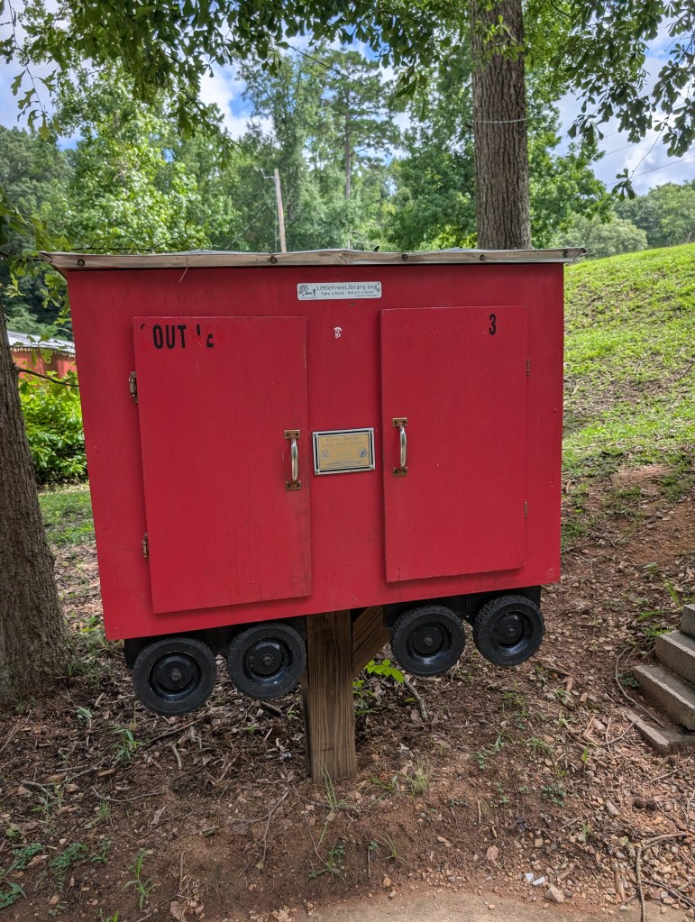 A Little Free Library that looks like a little red caboose with four black wheels at the bottom.
