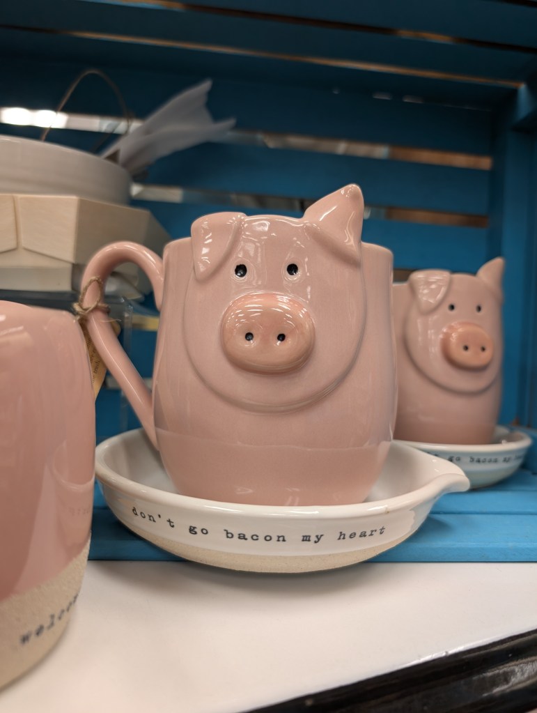 A ceramic mug in the shape of a cute pink pig. It rests in a white saucer that says "don't go bacon my heart".