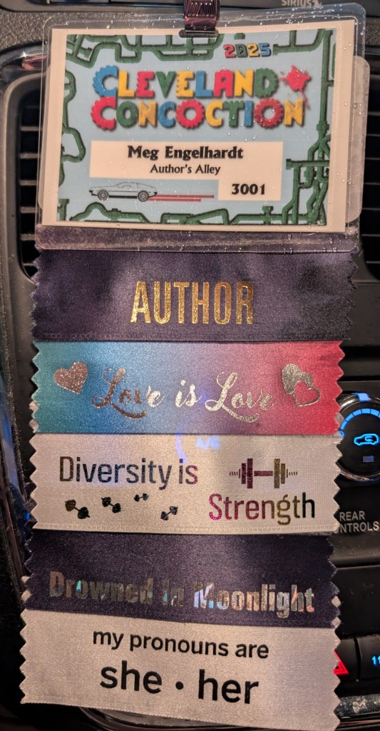 A badge that reads "2025 Cleveland Concoction - Meg Engelhardt - Author's Alley". It has ribbons attached that read: Author, Love is Love, Diversity is Strength, Drowned in Moonlight, My pronouns are she*her.