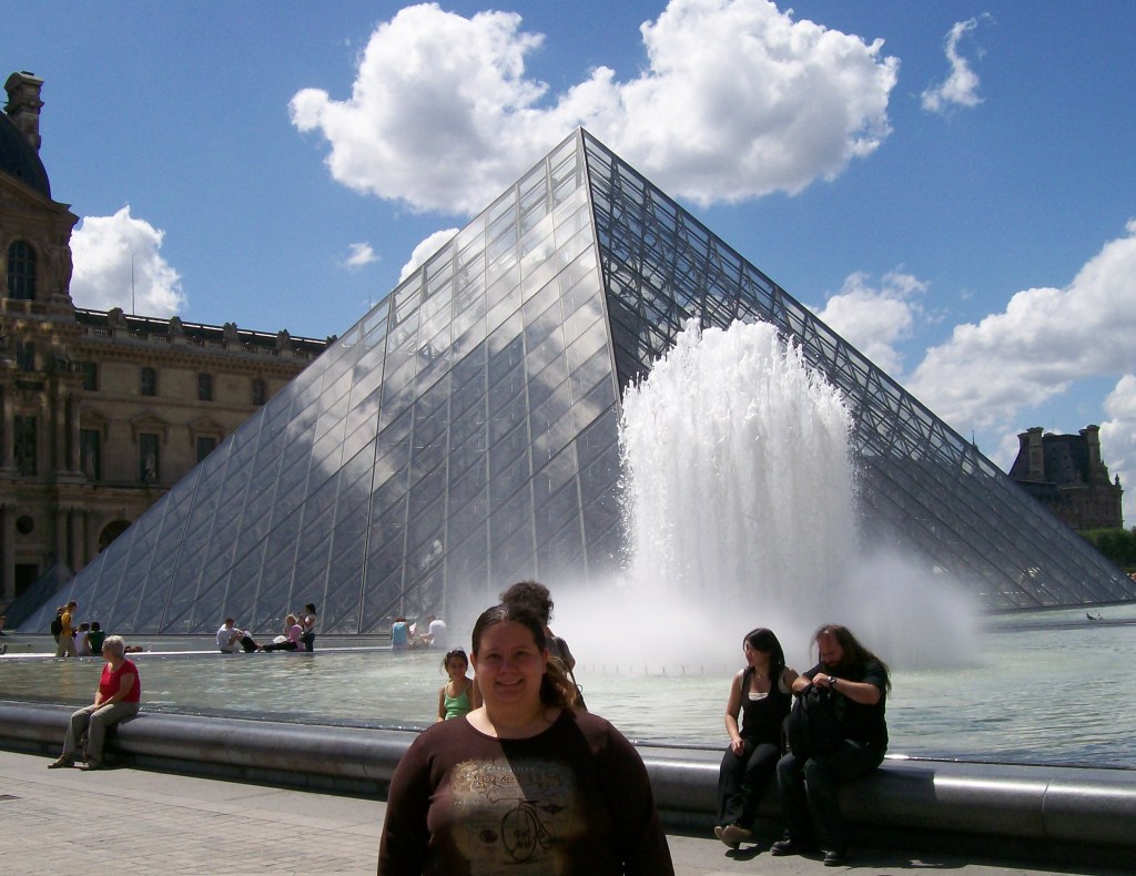 A round-faced person in front of a giant glass pyramid with fountains between. The Louvre has a pretty cool entrance. 