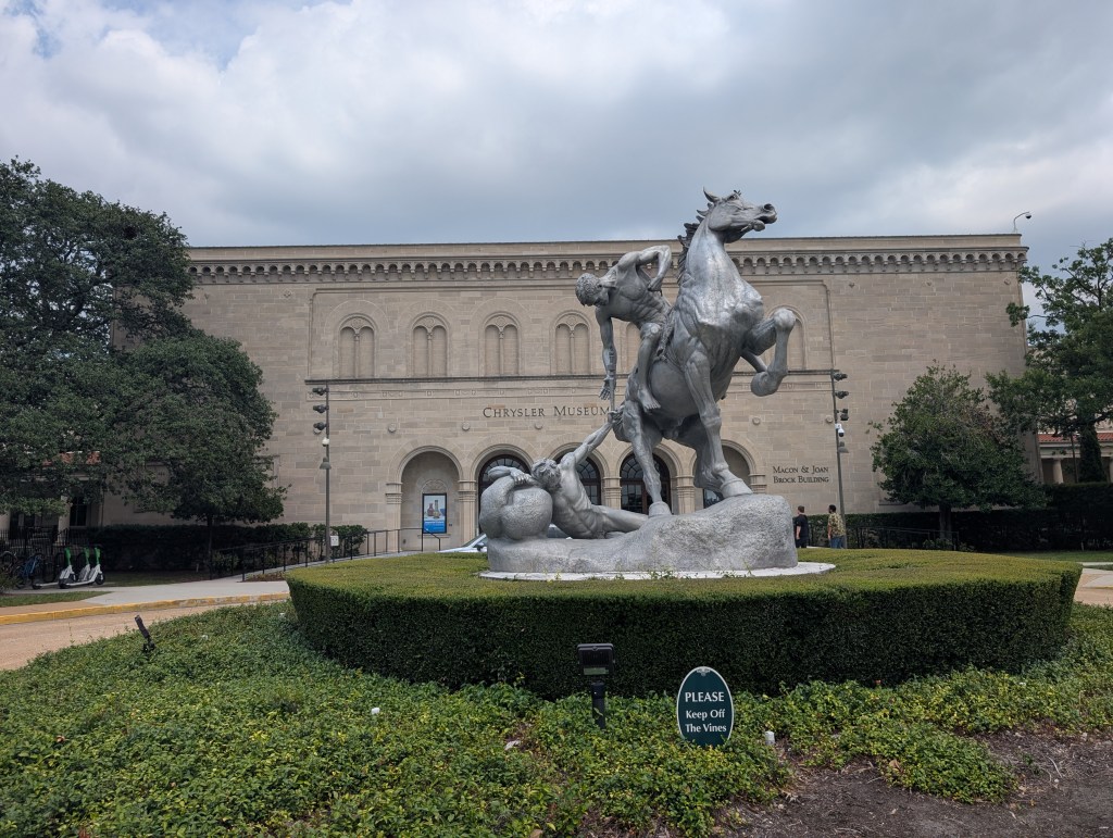 A large statue of a naked man on a rearing horse reaching down for a naked man on the ground, with a large museum front in the back. There are always lots of naked people in art museums. 