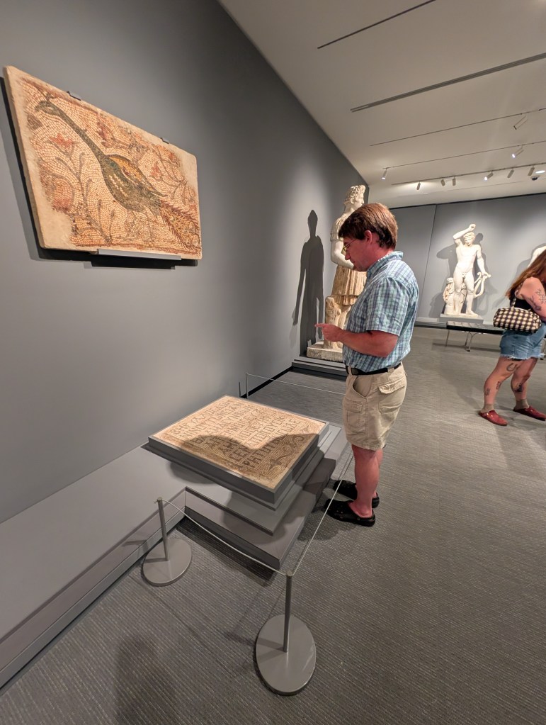 A middle-aged white man in khaki shorts and a blue button-up reading from a large square with ancient Greek writing on it. 