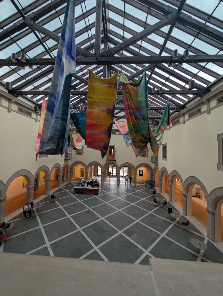 The entry to the art museum. It has large panes of glass as the roof and the girders are hung with several long banners that resemble hand-woven and hand-colored cloth. It is, in the manner of art museums, very liminal. 
